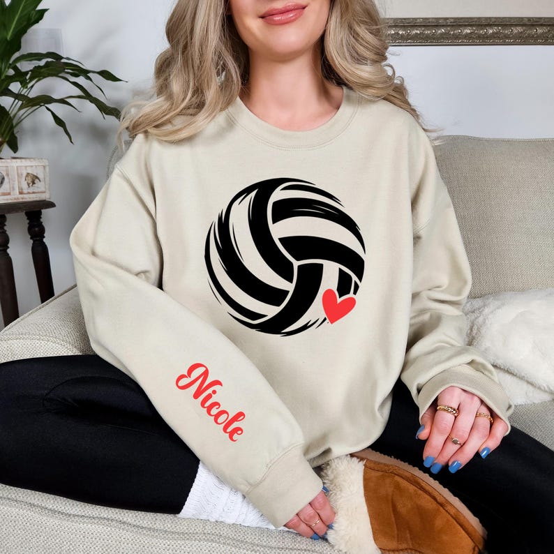 May include: Beige sweatshirt featuring a black and white volleyball design with a red heart accent. The sleeve displays the name "Nicole" in red script. The sweatshirt is a casual, comfortable garment.