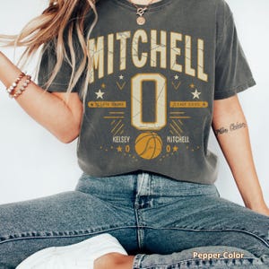 May include: A gray t-shirt with the name "MITCHELL" in large, distressed lettering. Below is a large number "0" with basketball stats and a basketball graphic. The shirt is a pepper color.