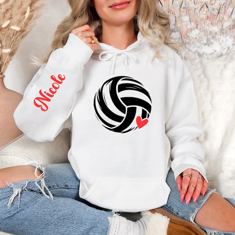 May include: White hoodie with a black and white volleyball graphic and a small red heart. The left sleeve has the name "Nicole" in red script. The hoodie has a drawstring hood and a front pocket.