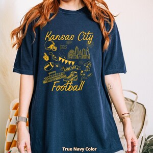 May include: A navy blue t-shirt with a yellow graphic design featuring the words "Kansas City Football." The design includes a football helmet, cityscape, football, and other football-related illustrations. The text "True Navy Color" is at the bottom.