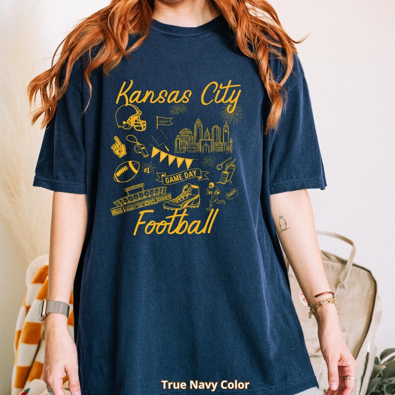 May include: A navy blue t-shirt with a yellow graphic design featuring the words "Kansas City Football." The design includes a football helmet, cityscape, football, and other football-related illustrations. The text "True Navy Color" is at the bottom.