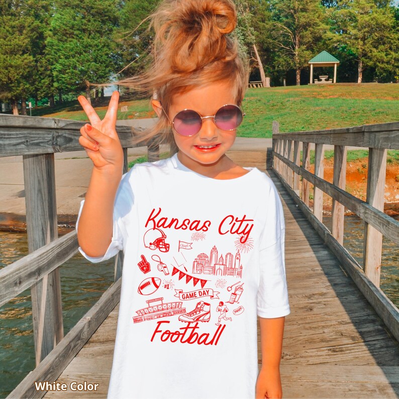 May include: White t-shirt with red "Kansas City" text and football-themed graphics. The shirt features a helmet, skyline, football, and the word "Football". The shirt is worn by a person wearing sunglasses.