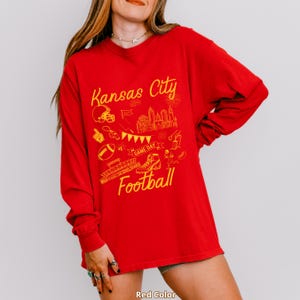 May include: A red long-sleeve shirt with a yellow graphic design featuring the words "Kansas City Football" and various football-related illustrations. The shirt is a solid red color.