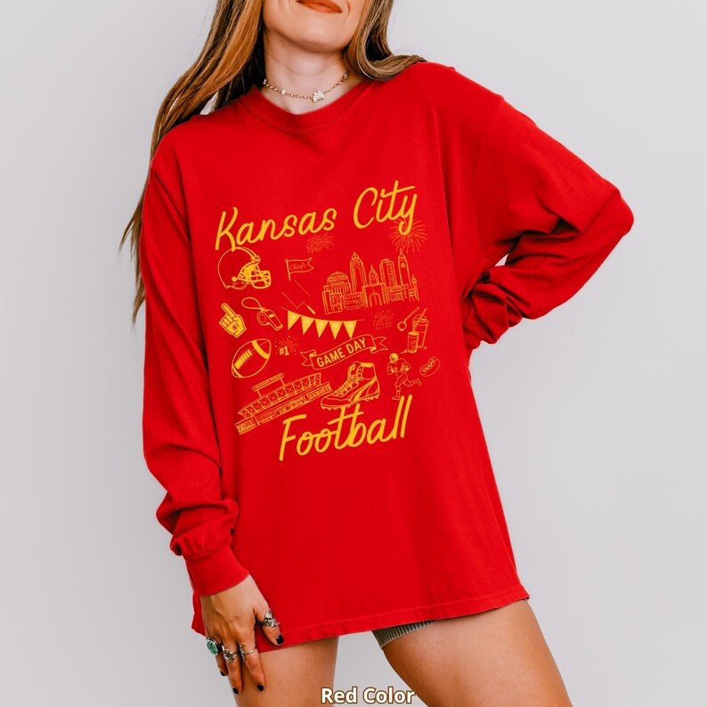 May include: A red long-sleeve shirt with a yellow graphic design featuring the words "Kansas City Football" and various football-related illustrations. The shirt is a solid red color.