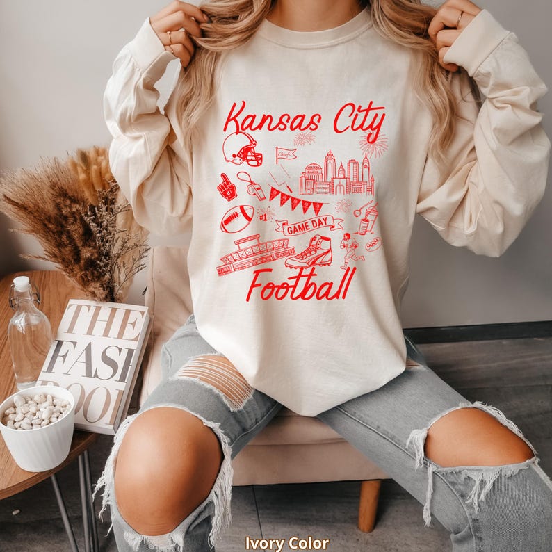 May include: Ivory-colored long-sleeve shirt with a red graphic design featuring the words "Kansas City Football" and various football-related illustrations, including a helmet, football, and cityscape.