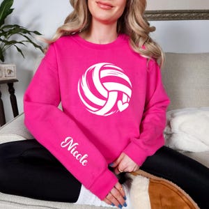 May include: A vibrant pink sweatshirt featuring a white volleyball design with a heart. The name "Nicole" is printed in white script on the left sleeve. The sweatshirt is a crew neck style.