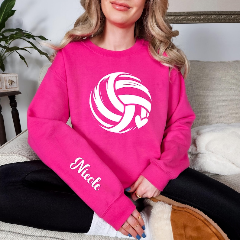May include: A vibrant pink sweatshirt featuring a white volleyball design with a heart. The name "Nicole" is printed in white script on the left sleeve. The sweatshirt is a crew neck style.