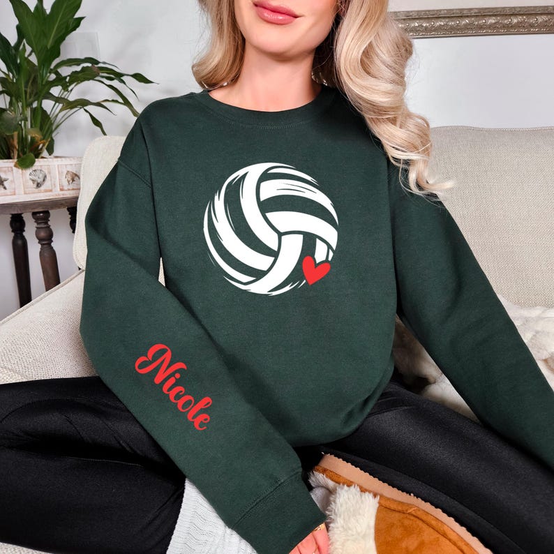 May include: Dark green sweatshirt featuring a white volleyball design with a red heart. The word "Nicole" is printed in red script on the left sleeve. The sweatshirt is a crew neck style.