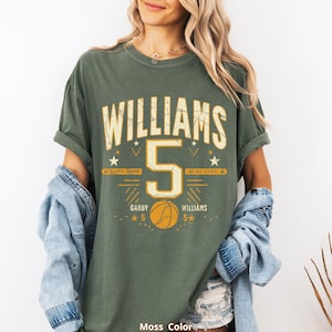 May include: Moss green t-shirt with the name "WILLIAMS" and the number "5" in a distressed, vintage style. The shirt features basketball-themed graphics, including a basketball and stars, with the text "GABBY 5" and "WILLIAMS 5".