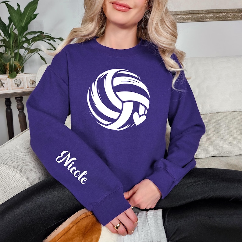 May include: A purple sweatshirt featuring a white volleyball design with a heart. The name "Nicole" is printed in white script on the left sleeve. The sweatshirt is a comfortable, casual style.