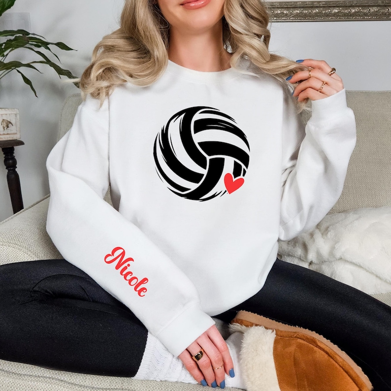 May include: White sweatshirt featuring a black and white volleyball design with a small red heart. The name "Nicole" is printed in red script on the left sleeve. The sweatshirt is a casual, comfortable style.