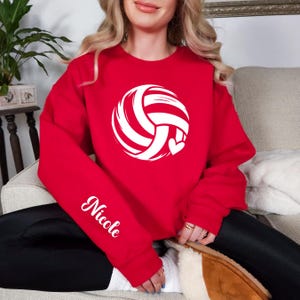 May include: A red sweatshirt featuring a white volleyball design with a heart. The name "Nicole" is printed in white script on the left sleeve. The sweatshirt is a solid red color.