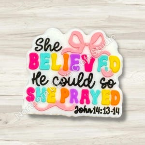 May include: A colorful, decorative piece with the words "She BELIEVED He could so SHE PRAYED" in various colors. A pink bow is at the top, and the text "John 14:13-14" is at the bottom. The piece is on a light wood grain background.