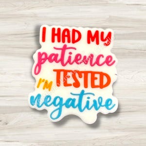 May include: A white, irregular-shaped charm with the text "I HAD MY patience I'M TESTED negative" in red, pink, orange, and blue. The charm has a glossy finish and is likely made of resin or a similar material.