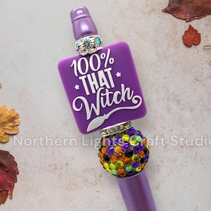 May include: A purple pen with a square charm that reads "100% THAT Witch" in white script. The pen features a jeweled band and a colorful, gem-studded sphere. The pen is decorated with a variety of colorful gems.