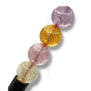 May include: A string of four glittery, spherical beads in shades of gold, pink, and purple. The beads are attached to a black base. The beads have a glittery texture and are arranged in a diagonal line.