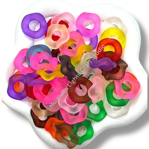 May include: A collection of colorful, frosted glass beads in various shapes and sizes, including circles and irregular forms. The beads are in shades of pink, purple, yellow, green, and brown, displayed in a white, flower-shaped dish. The text "Northern Lights Craft Studio" is visible.