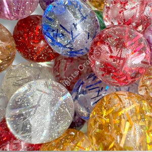 May include: Close-up of several clear, spherical resin beads filled with glitter and colorful metallic accents. The beads are in shades of red, blue, gold, and silver. The beads are scattered, creating a sparkling, festive display.