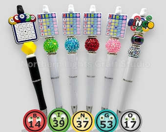 May include: Six pens featuring bingo-themed toppers with grid cards or colorful bingo balls. The pens have white or black barrels accented with sparkling rhinestone beads in green, blue, red, and pink. Five circular bingo ball icons with numbers 14, 39, 37, 53, and 17 are displayed at the bottom.
