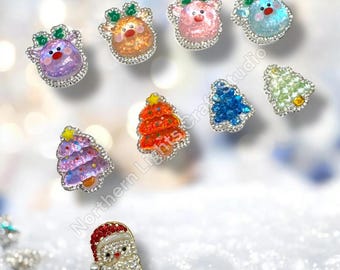 May include: A collection of small, decorative holiday-themed charms featuring glittery reindeer heads in purple, orange, pink, and blue, alongside colorful Christmas tree shapes and a Santa Claus face adorned with clear rhinestones.