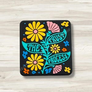 May include: Black rectangular magnet with a floral design and the words "TRUST THE PROCESS" in teal. The flowers are yellow, pink, and orange, with teal leaves and accents. The magnet is a decorative item.