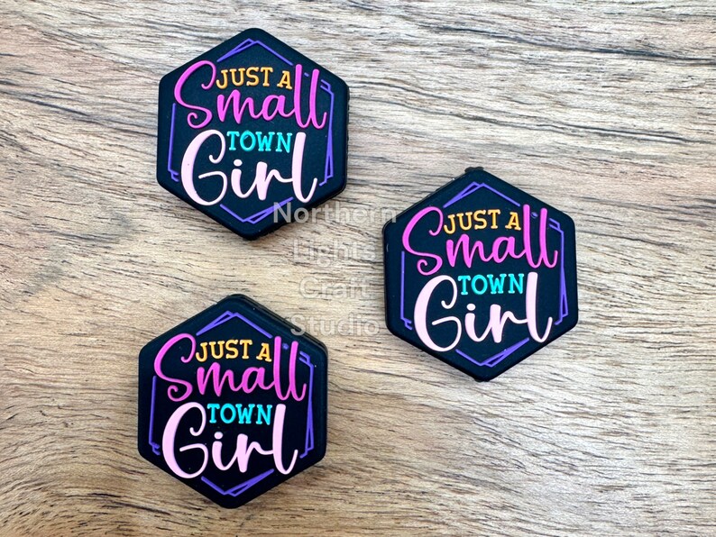 May include: Three black hexagonal charms with the text "Just a Small Town Girl" in colorful letters. The charms are on a woodgrain background.