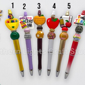 May include: Six decorative pens with various toppers, including a crayon box, a sign that says "getting my CRAY ON!", an apple, and a coffee cup. The pens have different colored barrels and are designed for teachers or students.