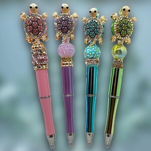 May include: Four decorative pens with turtle toppers. Each pen features a different color scheme: pink, purple, teal, and green. The turtle toppers are embellished with rhinestones. The pens have a silver-colored tip and a decorative band near the top.