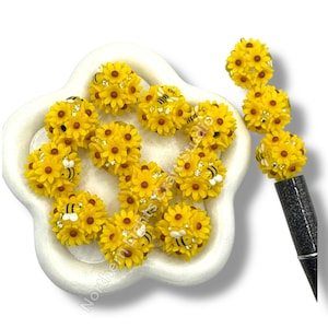 May include: A collection of yellow sunflower-shaped embellishments with brown centers and small rhinestone accents. Some embellishments feature a small bee design. The embellishments are arranged on a white, flower-shaped dish and a pen.