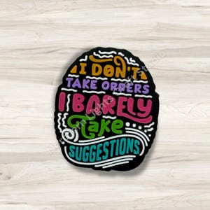 May include: Black, oval-shaped sign with the text "I DON'T TAKE ORDERS I BARELY Take SUGGESTIONS" in various colors. The text is in a whimsical, handwritten style. The background is a light wood grain.
