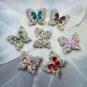 May include: Seven silver-toned butterfly-shaped embellishments featuring central teardrop-shaped crystals in blue, iridescent, green, clear, pink, champagne, and red, surrounded by smaller clear rhinestones on the wings.
