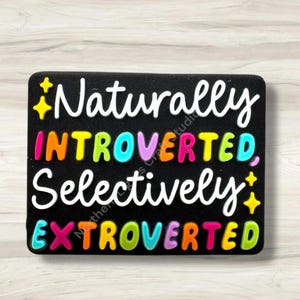 May include: A black rectangular charm with the words "Naturally Introverted, Selectively Extroverted" in white, and colorful letters. Yellow star accents are on the top left and bottom right.