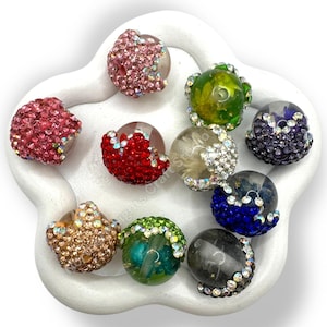 May include: A collection of colorful, decorative beads with crystal and rhinestone embellishments. The beads are in various colors, including pink, red, green, and blue, and are arranged in a white, flower-shaped dish. Each bead has a clear, rounded center.