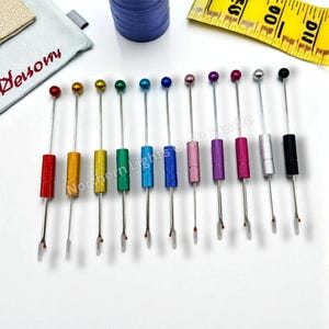 May include: A collection of colorful seam rippers with metallic handles and ball tops. Each ripper has a different color handle, including red, yellow, green, blue, pink, purple, and silver. The seam rippers are arranged in a row on a white surface.