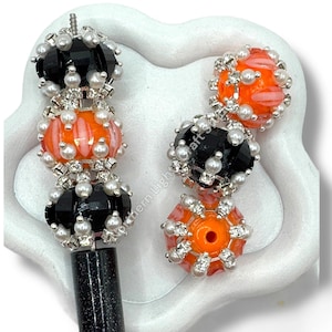 May include: Two decorative pens with black, orange, and silver beaded designs. The pens feature faceted black beads, orange glass beads, and small white pearl accents. The pen's barrel is black.