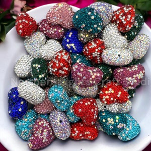 May include: A collection of heart-shaped rhinestone beads in various colors, including red, blue, green, pink, and silver. The beads are arranged in a bowl and are ready to be used for crafting projects.