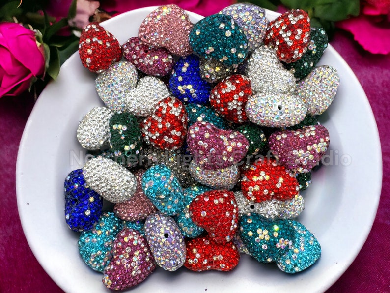 May include: A collection of heart-shaped rhinestone beads in various colors, including red, blue, green, pink, and silver. The beads are arranged in a bowl and are ready to be used for crafting projects.