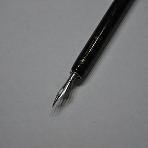 Może przedstawiać: Czarny długopis wieczny ze złotymi detalami i srebrną stalówką. Pióro jest ustawione po przekątnej na białej powierzchni, podkreślając jego elegancki design. Szczegółowość stalówki sugeruje kaligrafię lub precyzyjne pisanie.