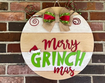 Może przedstawiać: Okrągły drewniany znak bożonarodzeniowy z napisem "Merry GRINCH mas" w kolorze czerwonym i zielonym. Znak posiada kokardę z juty z czerwoną i zieloną wstążką w kropki oraz dekoracyjne zielone i czerwone akcenty.