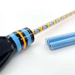 May include: A black glass wand with blue, orange, and black bands. The wand's shaft has a repeating pattern of blue and orange diamond shapes. Two light blue glass tubes are next to the wand.