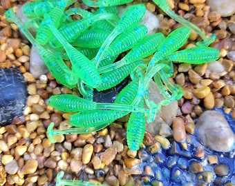 May include: A pile of green and blue fishing lures with a speckled finish. The lures are arranged on a bed of small brown and tan pebbles.