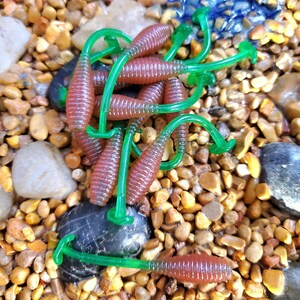 May include: A group of green and brown plastic fishing lures with a curly tail. The lures are arranged on a bed of small brown and yellow stones.