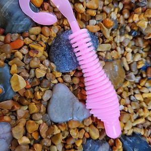 May include: Pink plastic fishing lure with a curly tail and a hook. The lure is on a bed of small rocks.