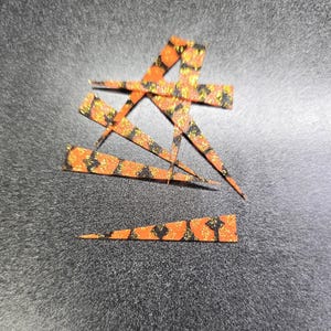 May include: A set of six orange, black, and gold glitter fish scales. The scales are shaped like a triangle with a pointed tip.