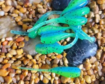 May include: A collection of soft plastic fishing lures featuring a dual-tone design of bright neon green and translucent blue. Each lure has a slender, elongated tail and a segmented body embedded with small, dark, reflective glitter particles.