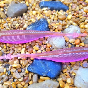 May include: Two pink plastic fishing lures on a bed of small brown and gray stones.