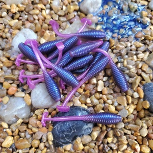 May include: A pile of purple and pink soft plastic fishing lures. The lures are shaped like worms and have a curved tail.