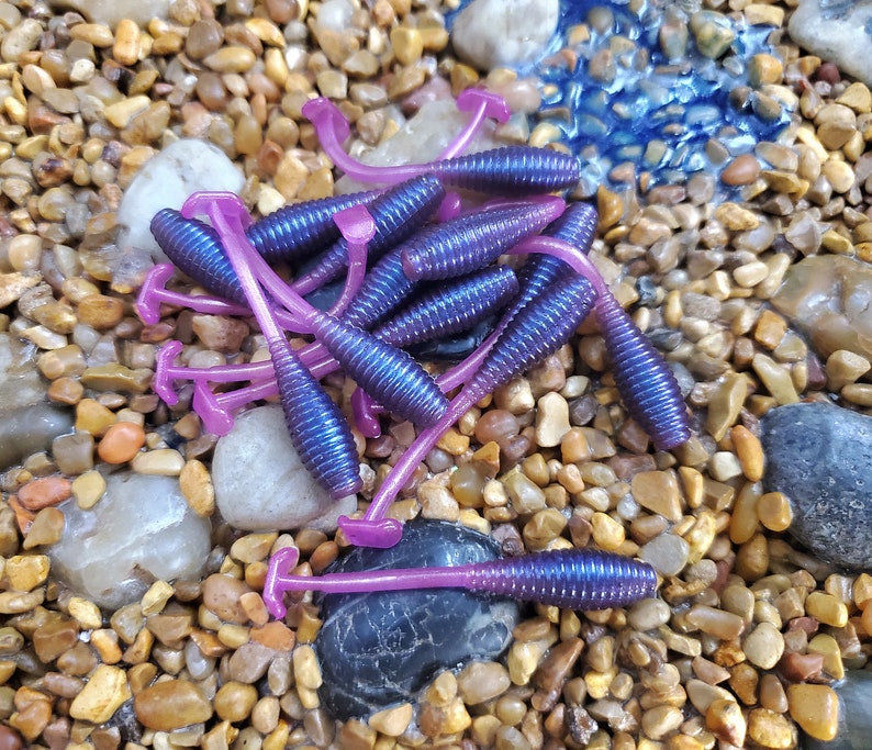 May include: A pile of purple and pink soft plastic fishing lures. The lures are shaped like worms and have a curved tail.