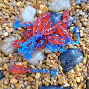 May include: A group of red and blue plastic fishing lures, resembling worms, are arranged on a bed of small brown and gray stones.