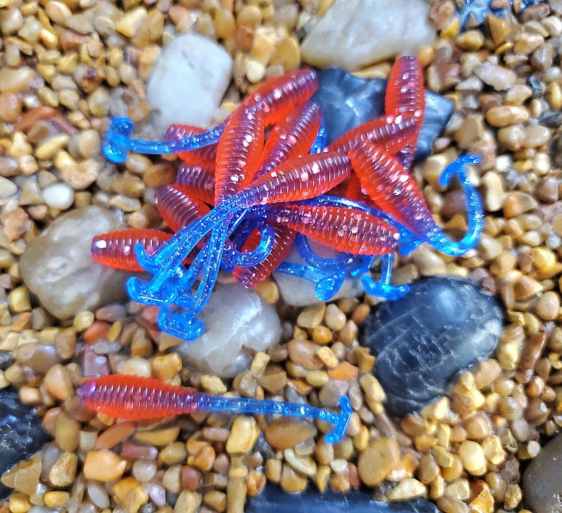 May include: A group of red and blue plastic fishing lures, resembling worms, are arranged on a bed of small brown and gray stones.